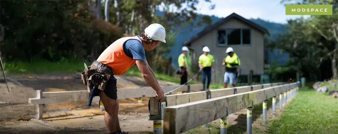 Featured Image_OnSiteBlog_January 2026_Modspace Builder measures timber framing on house piles at a building site, showing site preparation work that affects the costs of a transportable home.